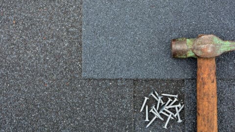 A hammer and a small pile of nails are laid across roofing shingles.