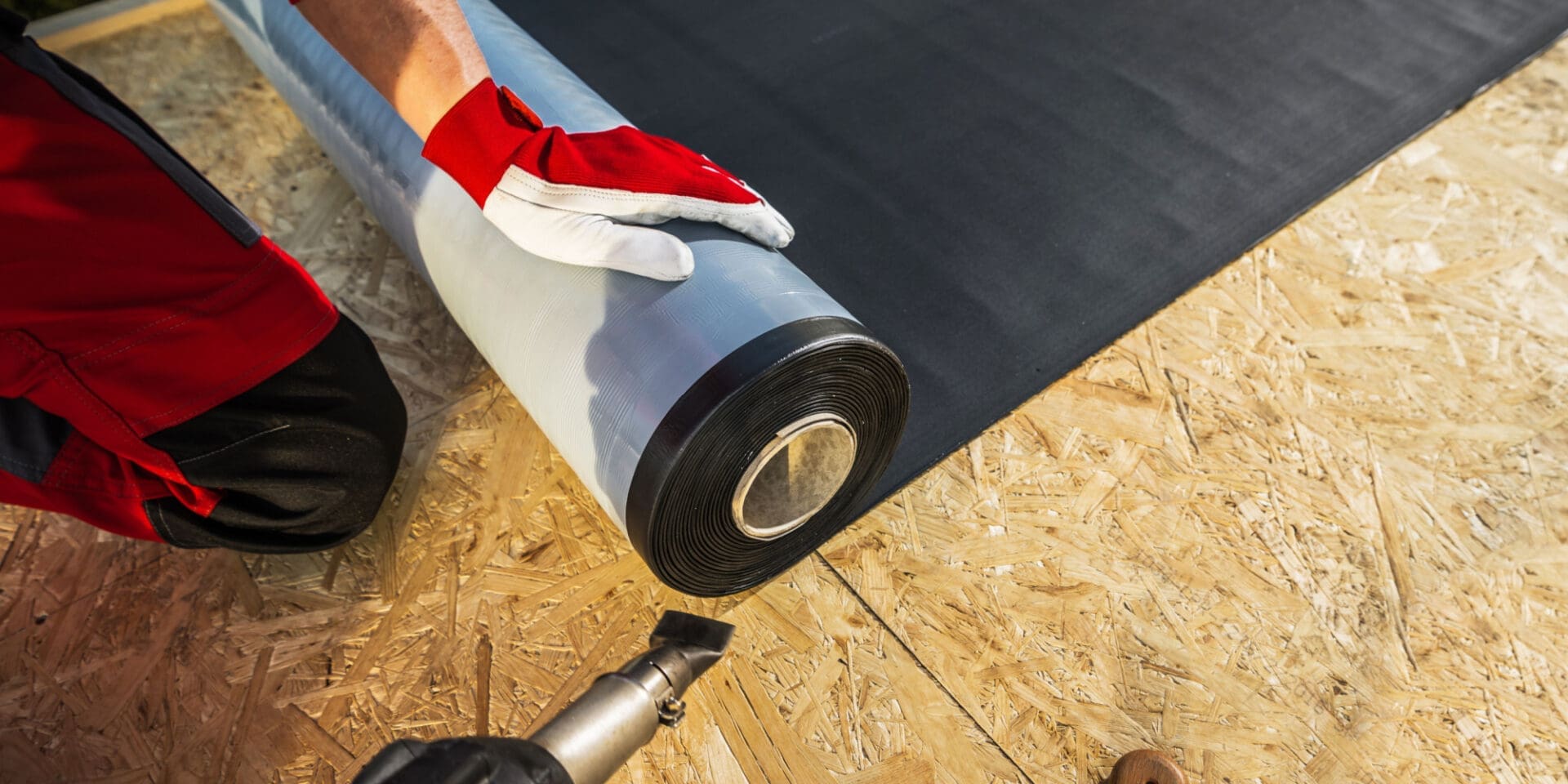 A roofer rolls out EPDM rubber roofing across the wooden decking of a roof.