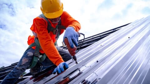 A roofer in a hard hat screws down metal panels onto a roof.