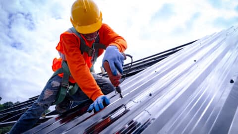 A man is on top of a steel roof installing it with a drill.