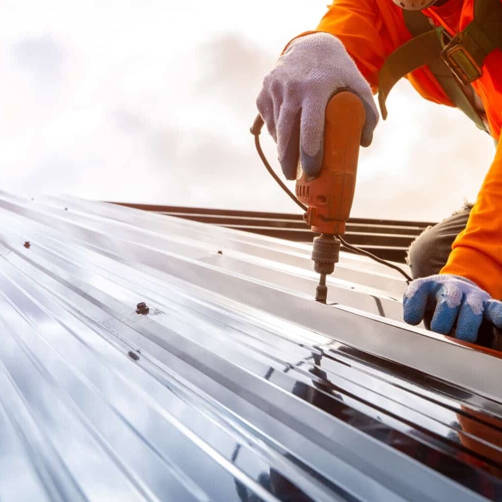 A roofer in a yellow shirt screws down corrugated metal sheets onto a roof.
